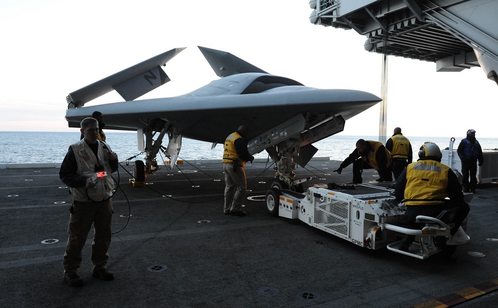 un X-47B les ailes repliées sur un porte-avions avec des personnes qui s'affairent autour
