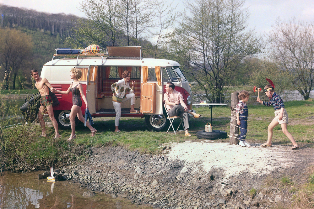 vu de profil droit, portes latérales ouvertes. Un couple en maillot de bain tient un matelas pneumatique et part se baigner. Une femme debout, un pied dans le véhicule parle avec un homme assis à une table. Une fillette déguisé en indienne joue avec son petit frère attaché à un tronc. Un voilier (jouet) flotte sur la rivière au premier plan