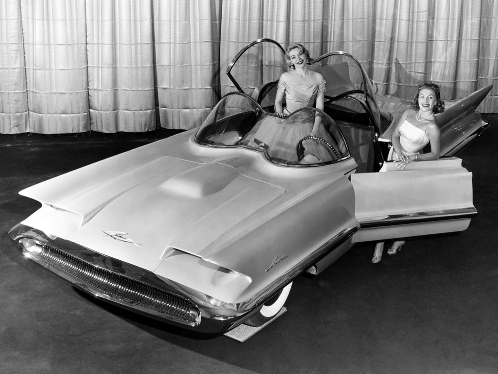 la Lincoln Futura 1955 vue de 3/4 avant gauche avec 1 femme debout dans l'habitacle montrant que la verrière supérieure s'ouvre et 1 autre femme derrière la portière ouverte